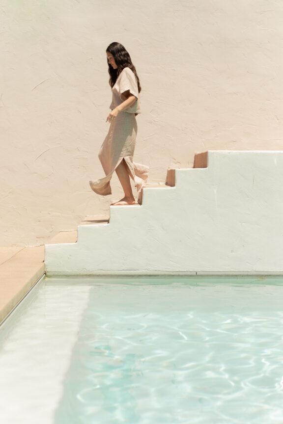 A woman in a light dress walks down white stairs beside a pool with clear water, against a beige textured wall.