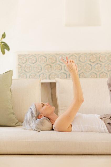 A woman with gray hair lies on a couch holding a smartphone above her, looking at the screen. The background features patterned tiles and neutral-colored cushions.