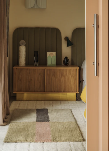 Bedroom with a partly open door revealing a wooden cabinet, modern decor items, striped rug, and an olive-green cushioned headboard.