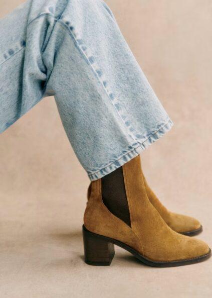 Person wearing light blue jeans and tan suede heeled ankle boots with dark elastic side panels, seated against a neutral background.