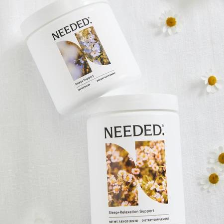 Two white containers labeled "NEEDED" on a white surface with small flowers; one is for stress support, the other for sleep-relaxation support.