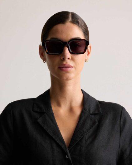 A woman wearing dark sunglasses and a black collared shirt poses against a plain light background.