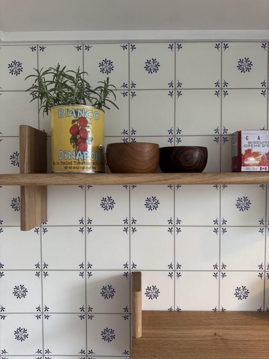 A wooden shelf holds a rosemary plant in a tomato can, two wooden bowls, and a box against a blue and white patterned tile wall.
