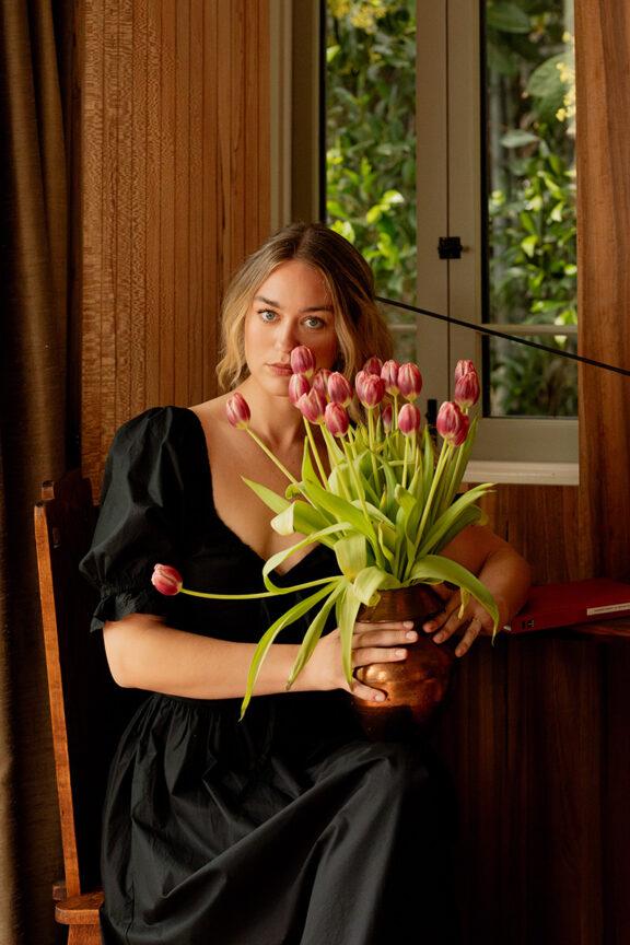 A woman in a black dress sits indoors, holding a brown vase filled with pink tulips, with a window and greenery in the background.
