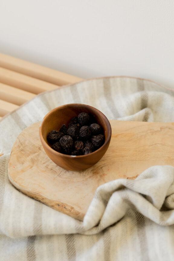 A wooden bowl filled with small, dark fruit sits on a wooden board atop a striped cloth.