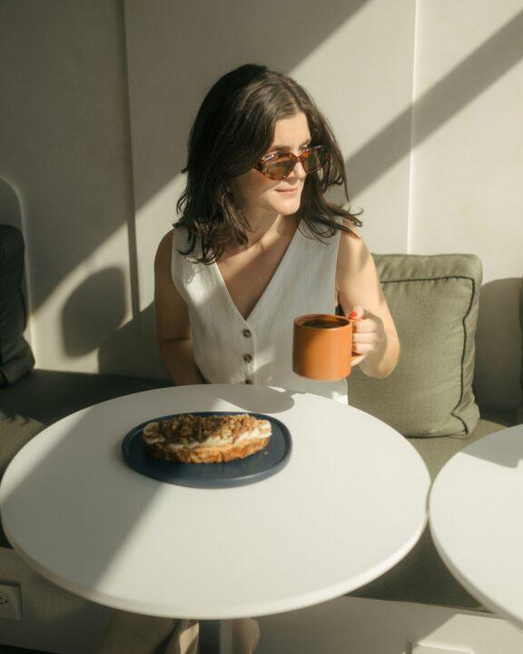 Woman in sunglasses sits at a round white table holding a mug of coffee, with a plate of pastry in front of her. Sunlight casts shadows across the scene.