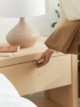 Person holding a brown sweater opens a wooden nightstand drawer beside a table lamp and book.