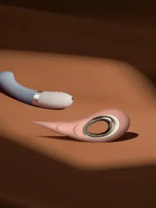Two modern sex toys, one blue and curved with a silver accent and one pink with a circular opening, placed on a brown, softly lit surface.