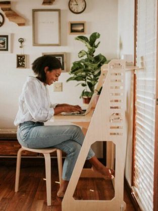 Woman sitting at a desk, using a laptop, in a well-lit room with wall art and plants.